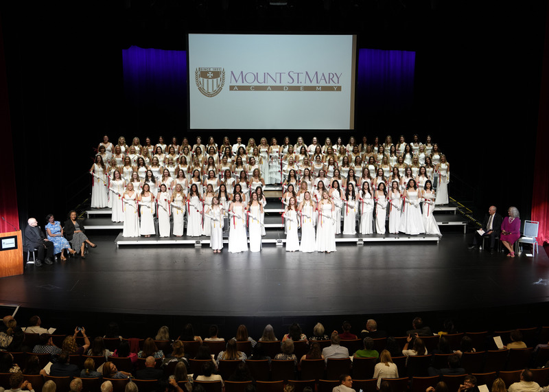 large group of graduates in long white gowns and white gloves standing on stage during their graduation ceremony