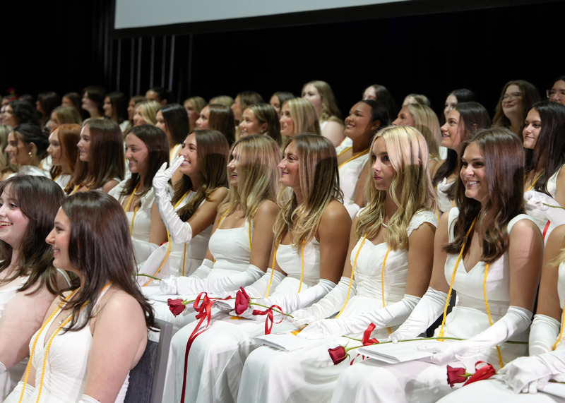 large group of young women wearing white gowns and white gloves, seated and smiling on stage during their graduation ceremony