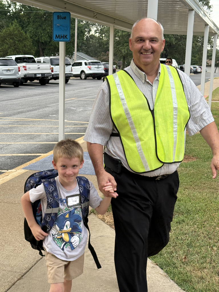 Kindergarten Principal MR. Roach wearing a traffic vest and holding the hand of a Kinder student. 