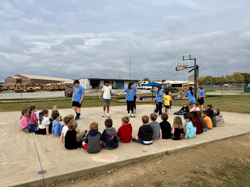 teens playing on basketball court with kindergarten students seated around them. 