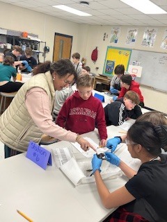 a teacher and middle school kids wearing gloves and goggles dissect a frog