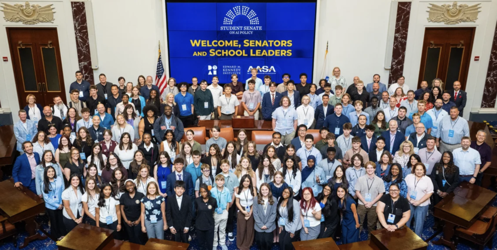 A gathering of students and teachers looking  toward the camera with a screen that displays "Student Senate on AI Policy, Welcome Senators and School Leaders."