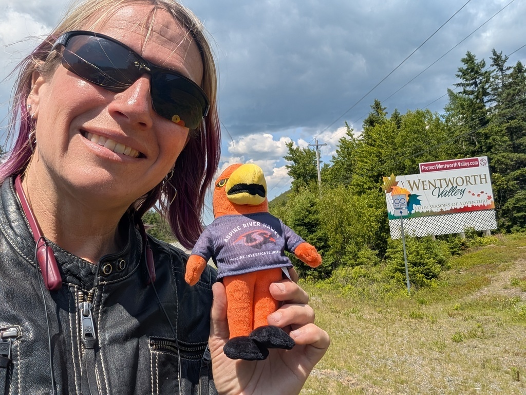 Teacher outside with River Hawk plushie in front of Wentworth Valley sign