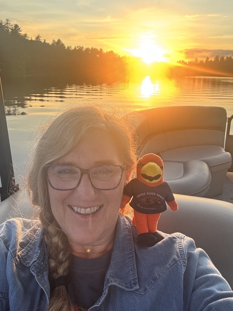 Teacher outside with River Hawk plushie in front of sunset