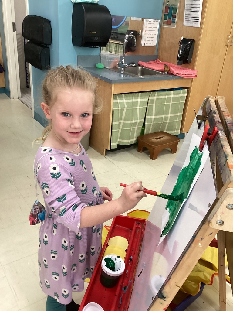 A girl in purple dress painting a green dot on a paper on a easel.