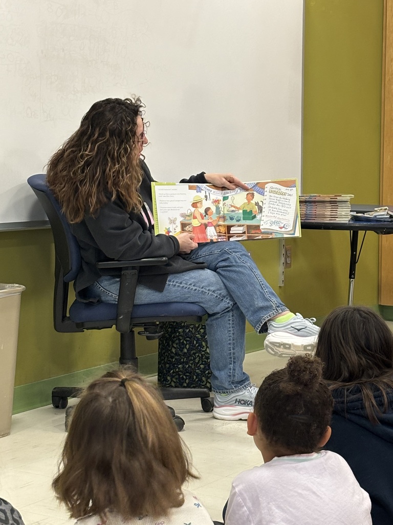 An adult woman reads a book to a group of students on the floor.