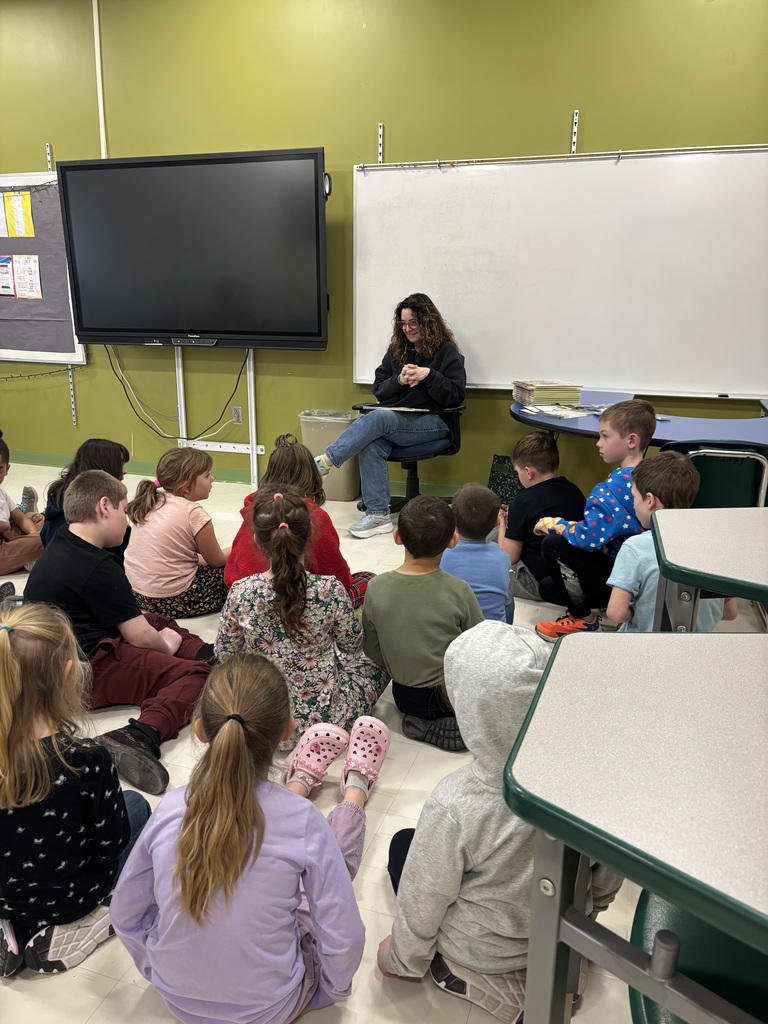 An adult woman reads a book to a group of students seated on the floor.