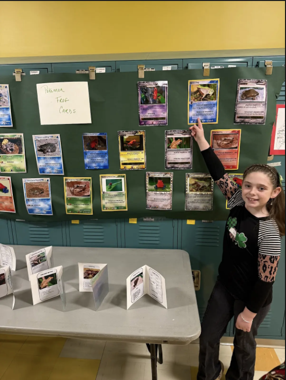 A girl points to a frog card on the wall.