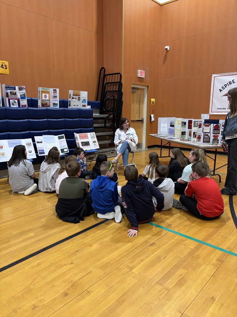 Students sit on floor looking at teacher reading a book.