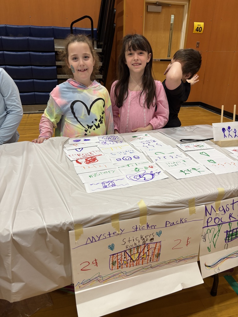 2 girls stand at a table selling stickers. 