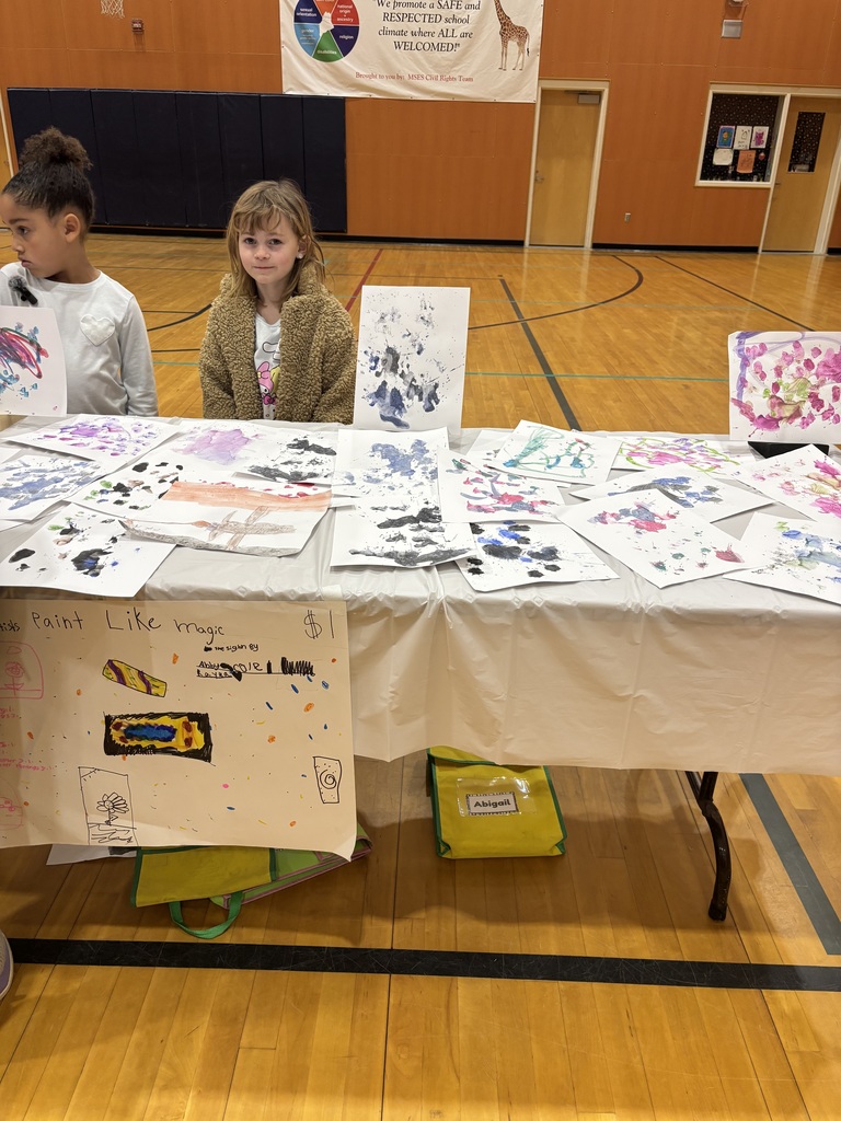 Two girls stand at a table selling paintings.