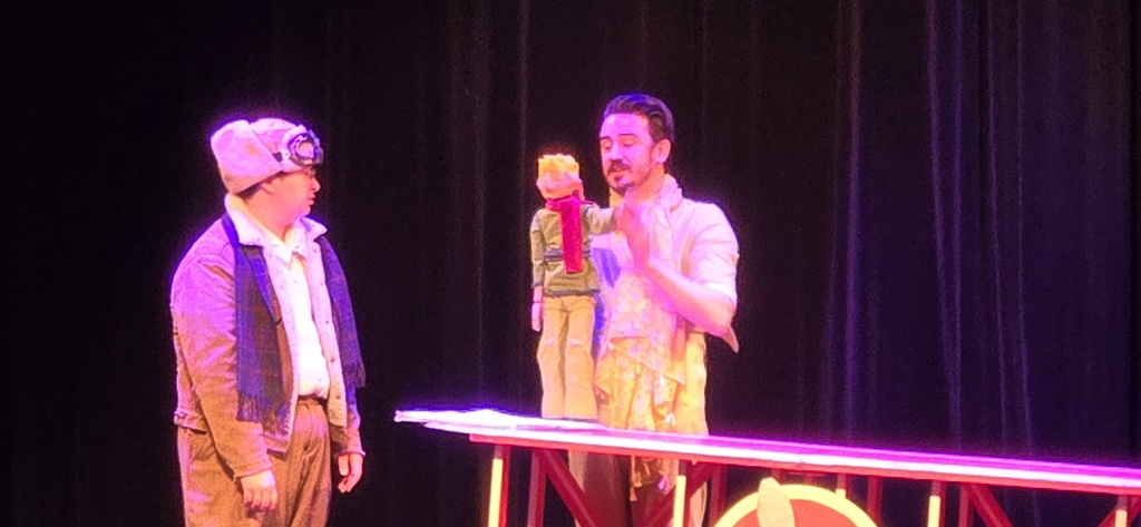 Two men look at a doll figure on stage.