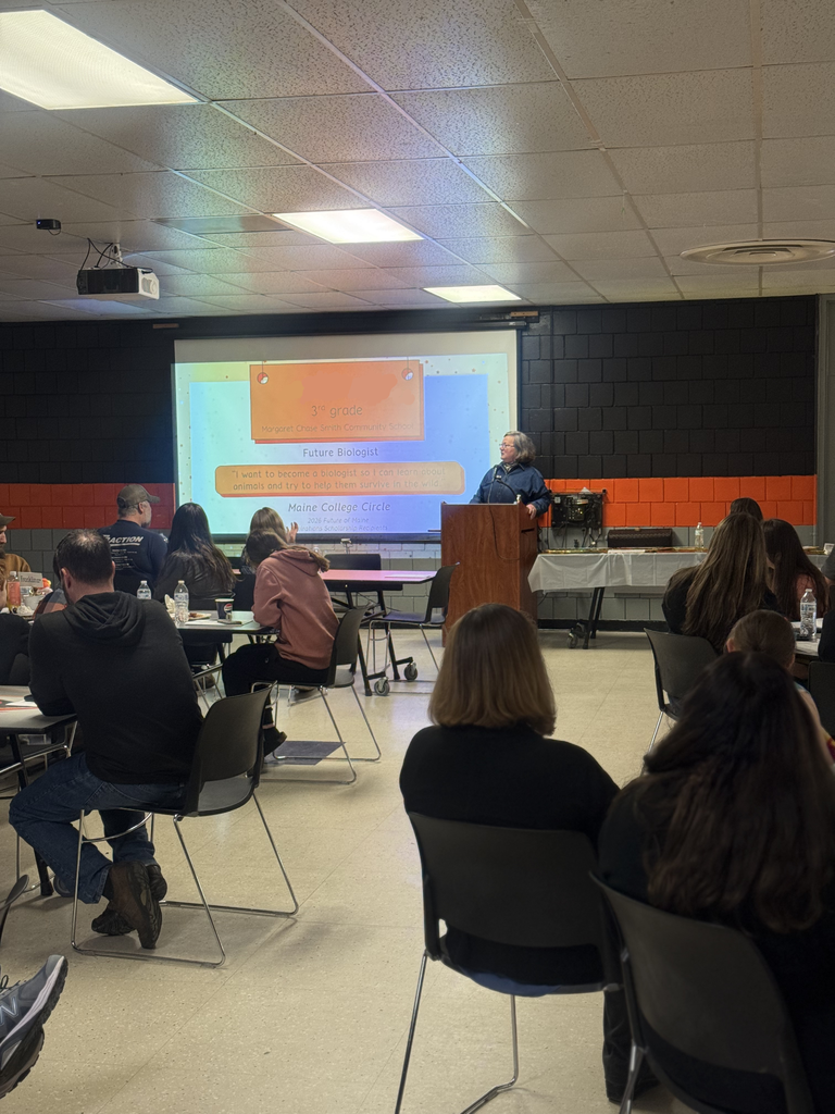 speaker in front of crowd in cafeteria. wall has a large screen with a slideshow highlighting student aspirations