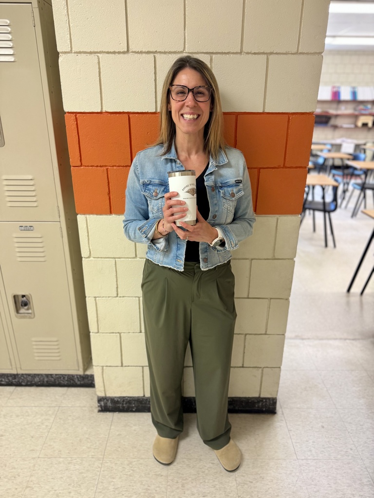 teacher holding cup and smilingin school hallway in between classroom doorway and lockers