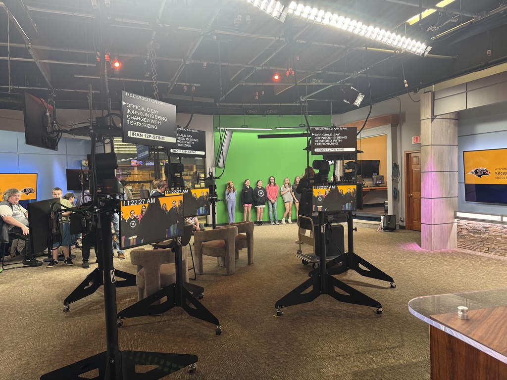 students in news filming room. cameras and tv screens in the foreground, students in front of greenscreen in background. 