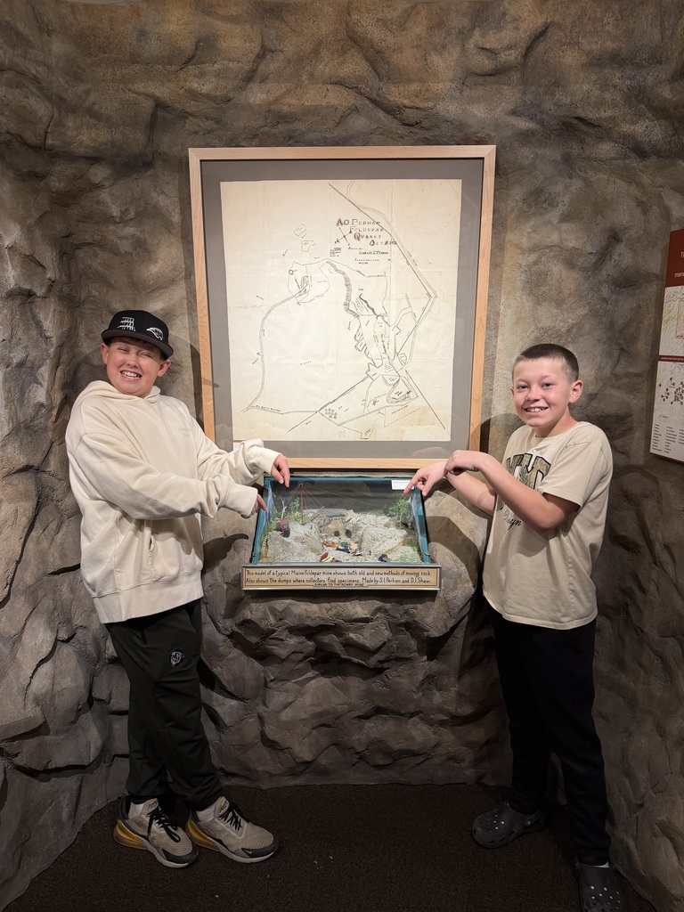 two middle school students in museum pointing to display case with gem samples, in front of map