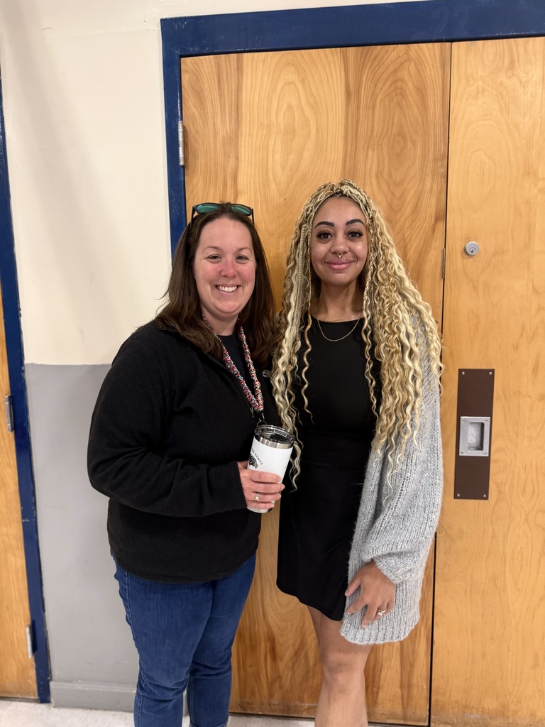 teachers smiling for photo, holding a white cup, in front of door in school hallway