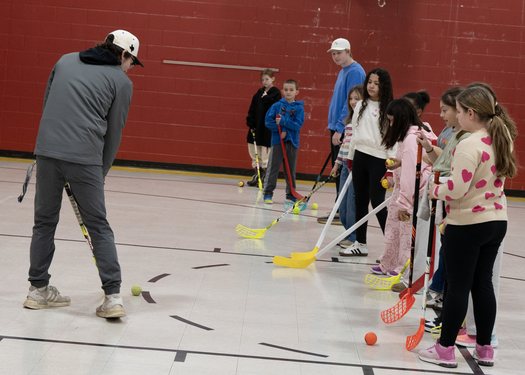 man showing line of students how to use a stick and ball for hockey