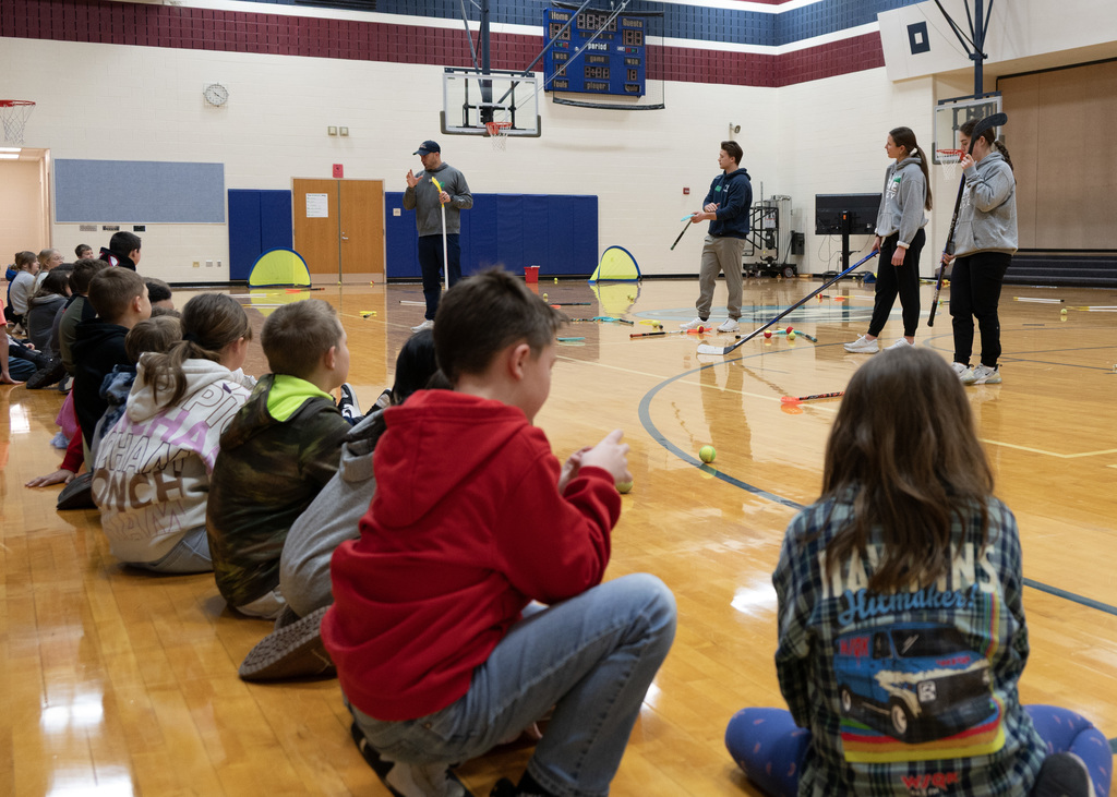 elementary students in a sitting in line being talked to by 4 hockey players
