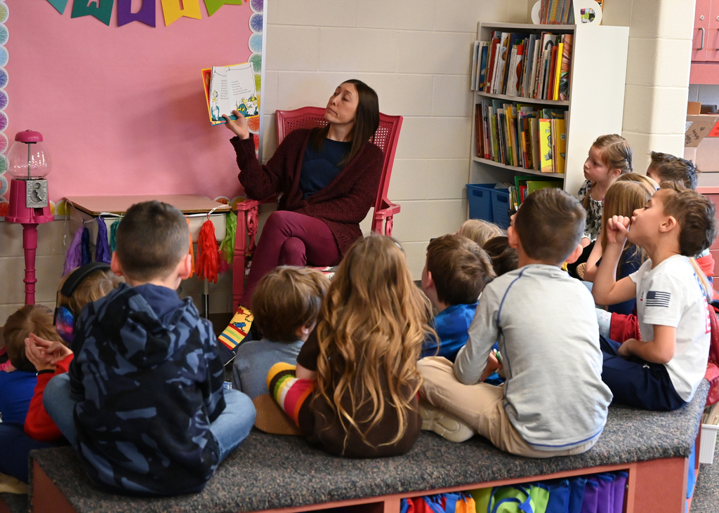 Teacher sitting in chair in front of students reading a book