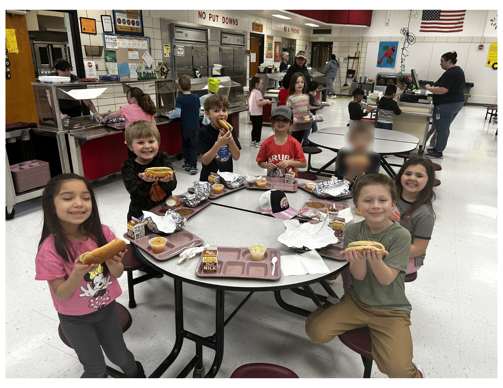 Childern sitting arounf a cafeteria table eating hot dogs