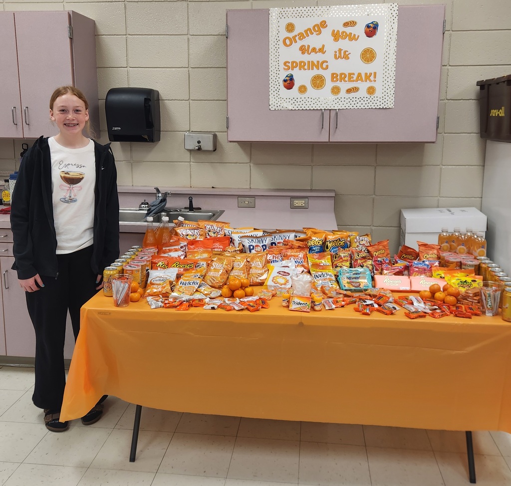 A girl standing next to a table filled with snacks and drinks all in packages colored orange with a sign that says orange you glad its spring break