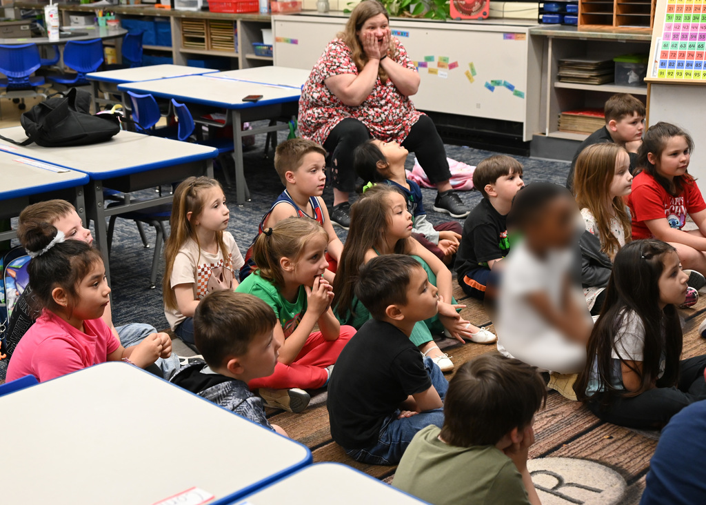 Childern sitting on floor listening to story reacting , teacher in back sitting reacting in surprise