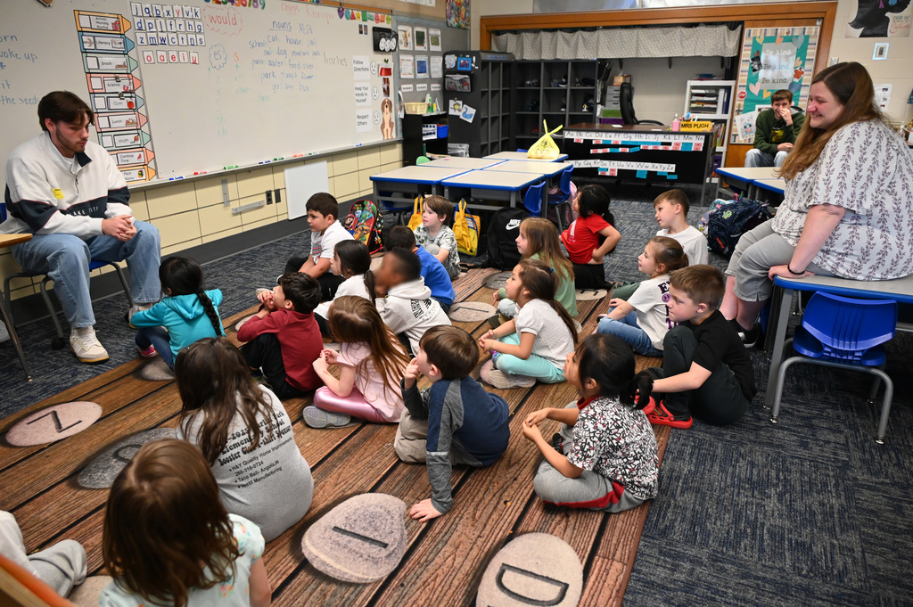 children sitting on floor listening to story being told by person in front of class as teacher looks on from the back