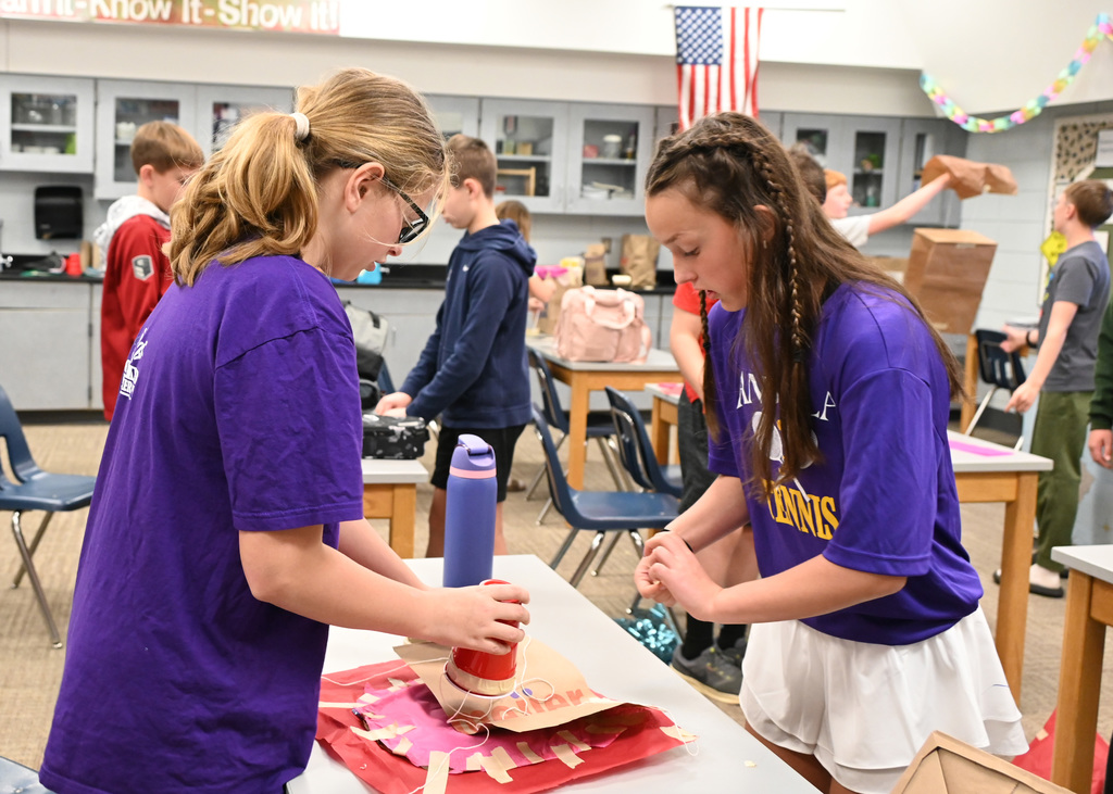 Two middle schooler working on a project with other childern in back also working on projects