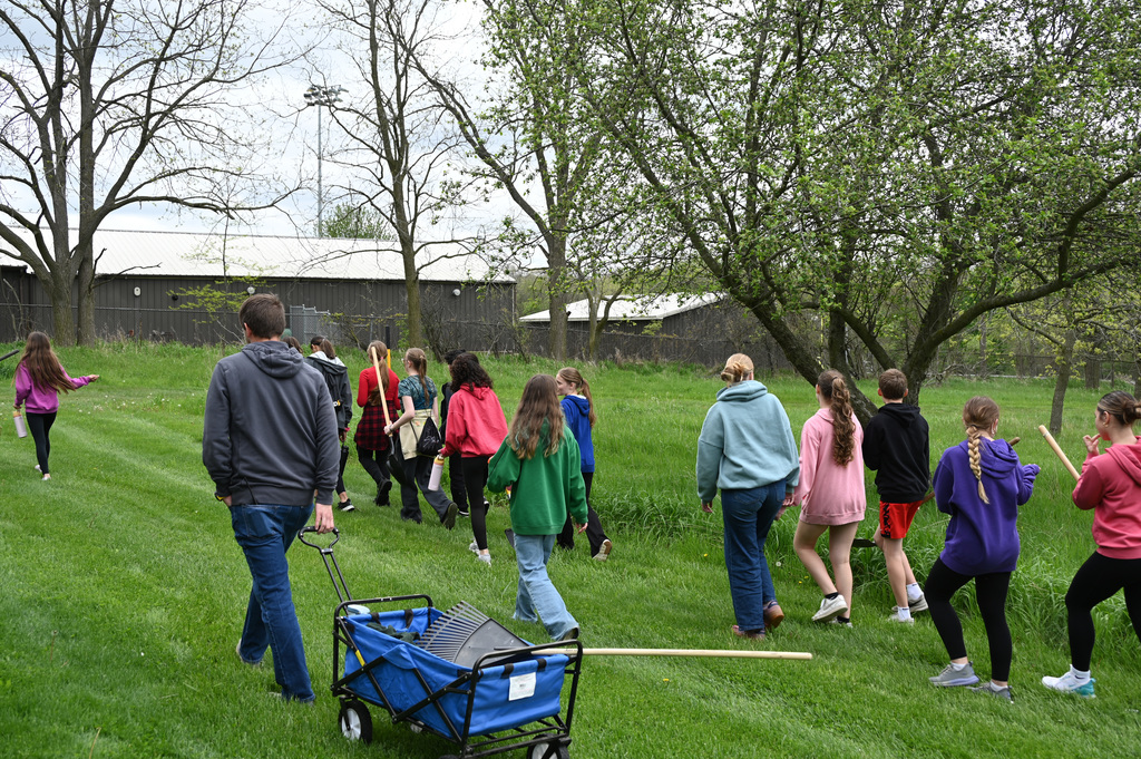 A class of middle school students walking in a line in a park carrying shovels and rakes