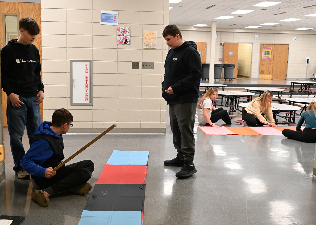 three boys measuring, with girls in back also mesuring long sheets of paper