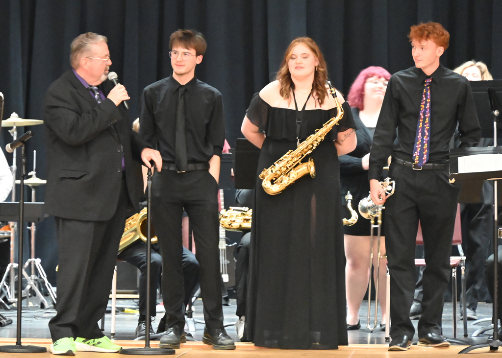 a man with microphone , teen with arms behind him, tean girl with saxaphone and boy with trumpet standing on a stage