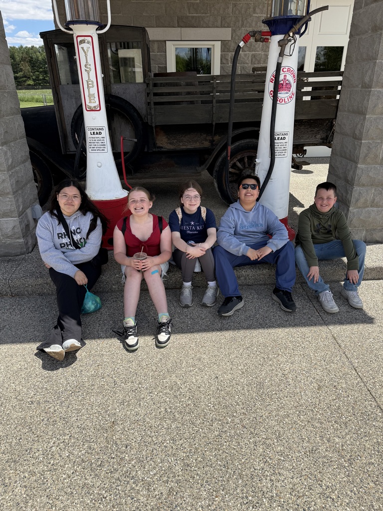five fithe graders sitting on ground in fornt of old car smiling