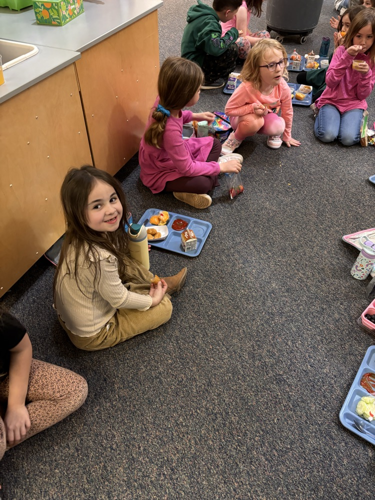 girls sitting on floor with her lunch tray smiling at camera, other children can be seiin sitting on floor behind her