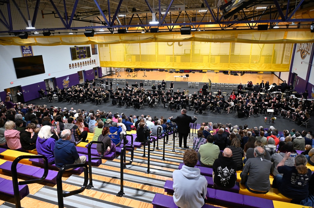 Gym filled with people a band fills the floor conductiors stand in bleachers leading