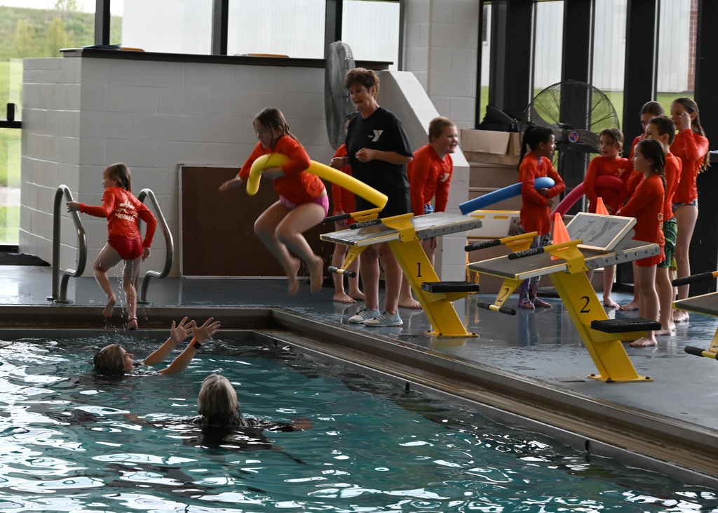 girl jumping in pool with noodle as other children wait to jump in also