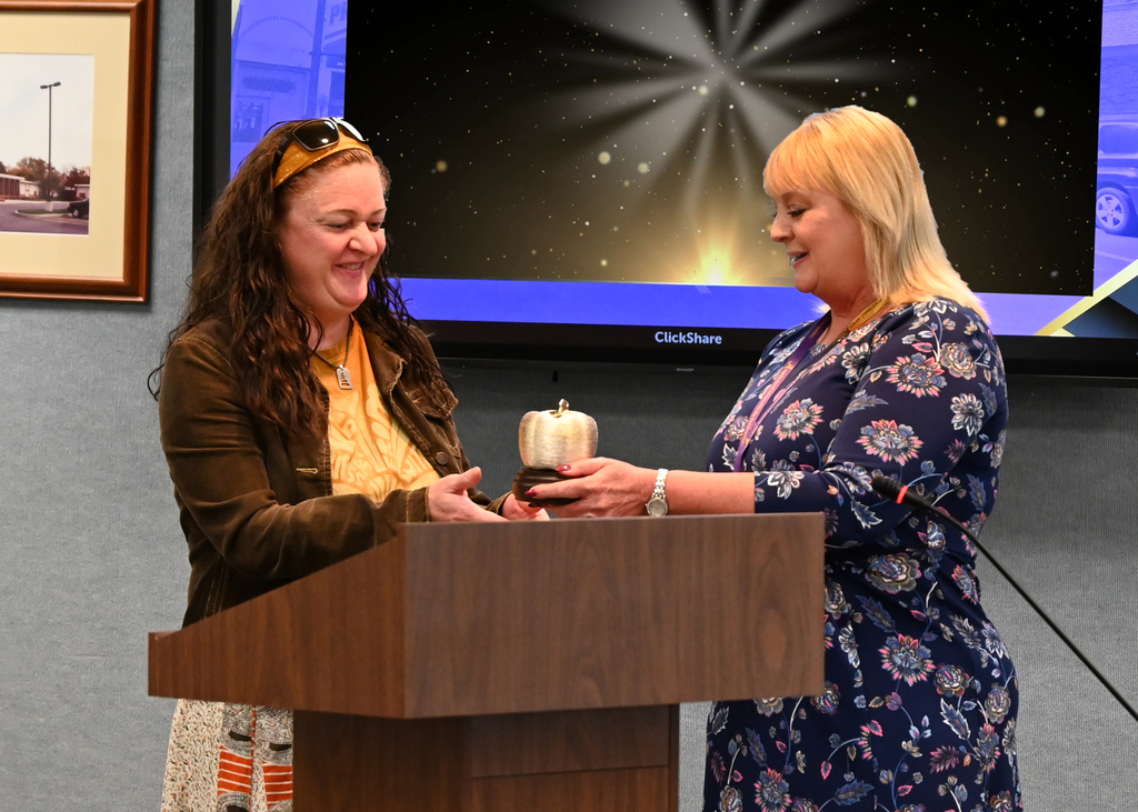 two women by a pedestal one is handing the other one a golden apple