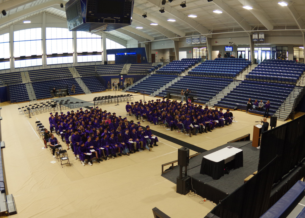 gymansiums with students in purple graduation gowns sitting in seats on floor