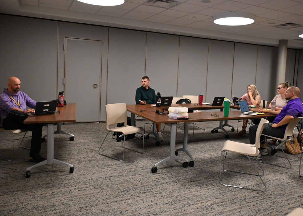 man at front of room at a desk with a compture talking with group of four people at working at another table