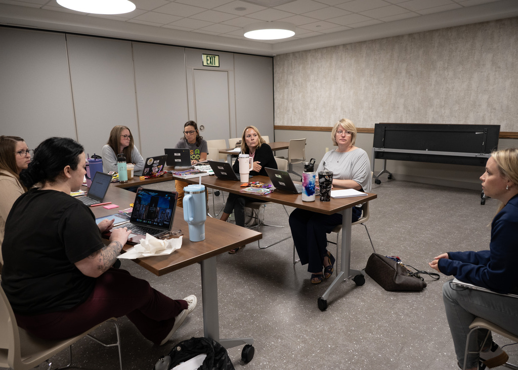six people sitting around a u shaped table on laptops with person in chair in front of U talking with them