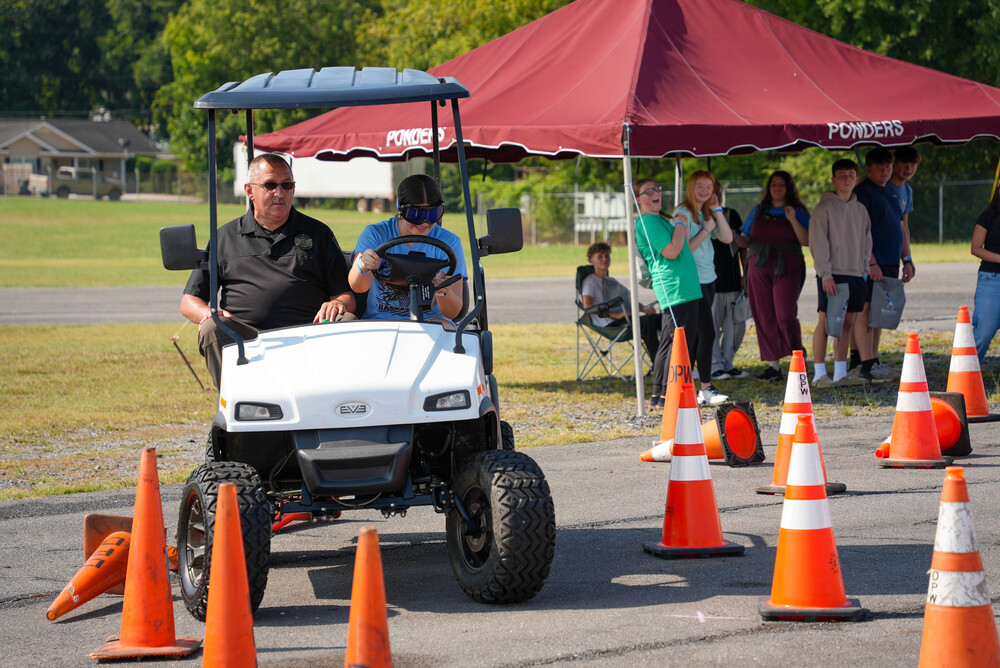 Girls drives golf cart with male officer in passenger seat and onlookers in background