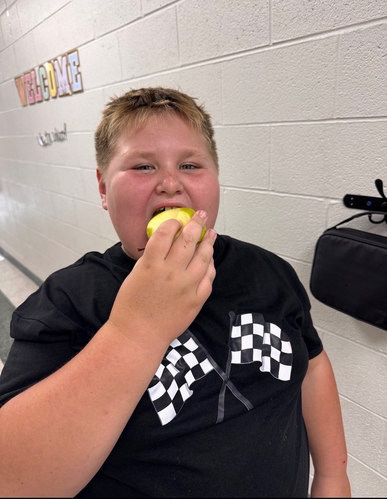 A person wearing a black t-shirt with a checkered pattern eats an apple in a hallway.
