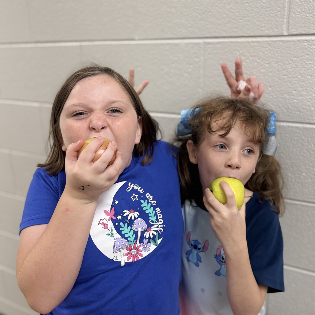 Two children eat apples while standing against a white wall. One wears a blue shirt with a graphic.