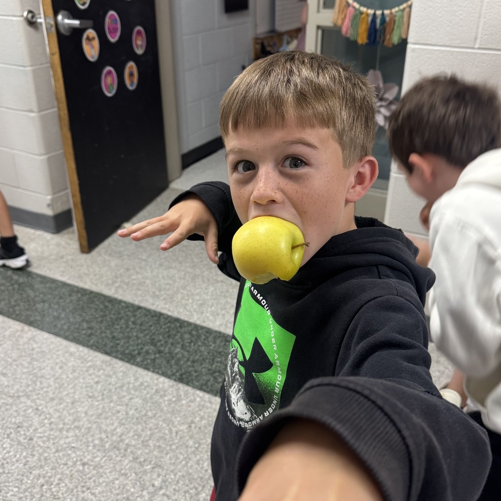 A boy holds an apple in his mouth in a hallway. Another child stands behind him, and a door is in the background.