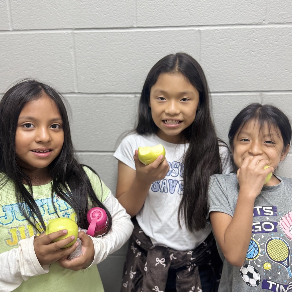 Three girls stand together. Each holds a lemon. The girl on the right eats it. White wall behind them.