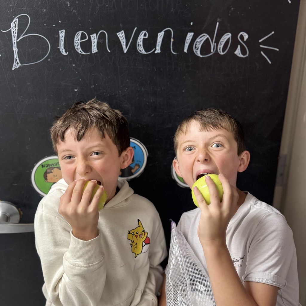 Two boys sit in front of a blackboard with "Bienvenidos" written on it, eating apples.