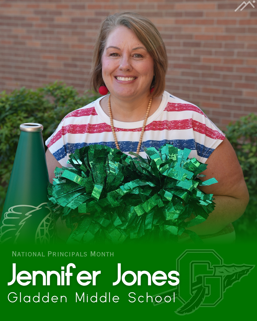 A smiling woman holds a pom-pom while standing in front of a brick wall and green bushes.