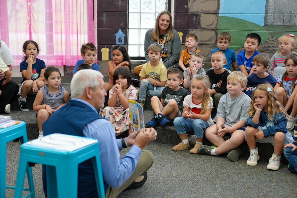 Group of children sitting on floor, listening to a man read aloud. Background includes a wall with a mural.