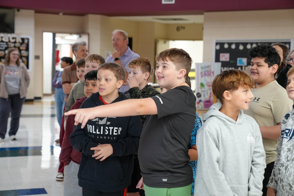 School children stand in line, with a boy in a black shirt pointing. Adults and more children are in the background.