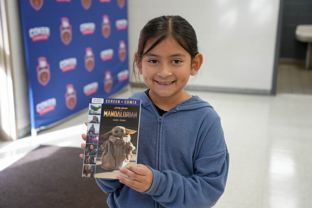 Girl holds book with image of Grogu, smiling. Behind her is a blue wall with logos.